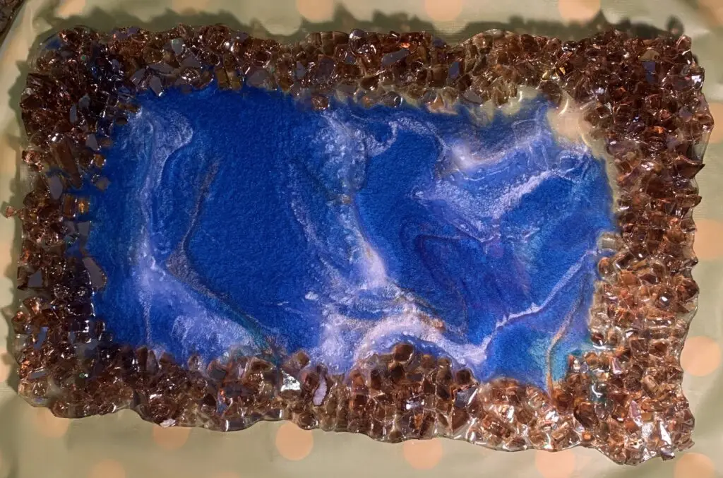 "Two Hearts" is a rectangular resin artwork with vibrant blue swirls resembling flowing water or marble. It is framed by amber-toned crystal-like fire glass, adding texture and contrast. The interplay of the fluid blue center and earthy border evokes natural elements such as rivers or oceans bordered by minerals.