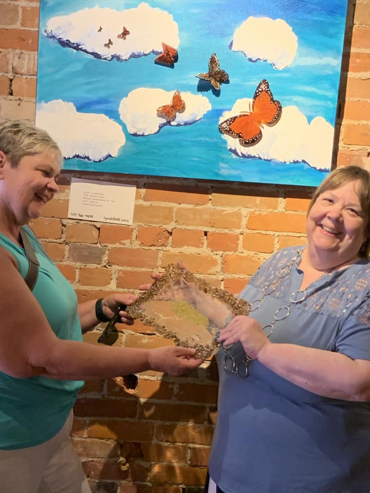 Karen Warner and Lynda Todd hold a resin tray standing in front of the piece Monarch Skies.