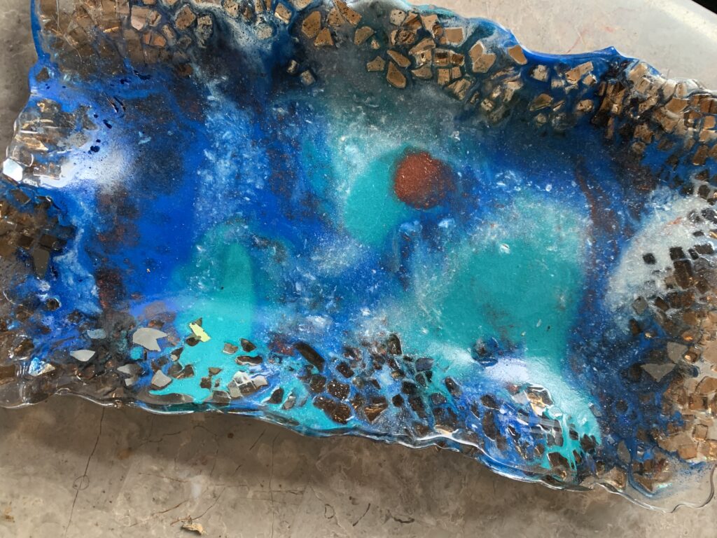 "Ripple of Celestial Waters" is an 8" x 12" resin artwork featuring vibrant blues and teals, with a striking red spot at its center. The surface has wavy, irregular textures, with glossy and matte finishes, reminiscent of an oceanic or cosmic landscape.